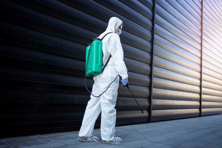 Person in white chemical protection suit doing disinfection and pest control with sprayer to kill insects and rodents.